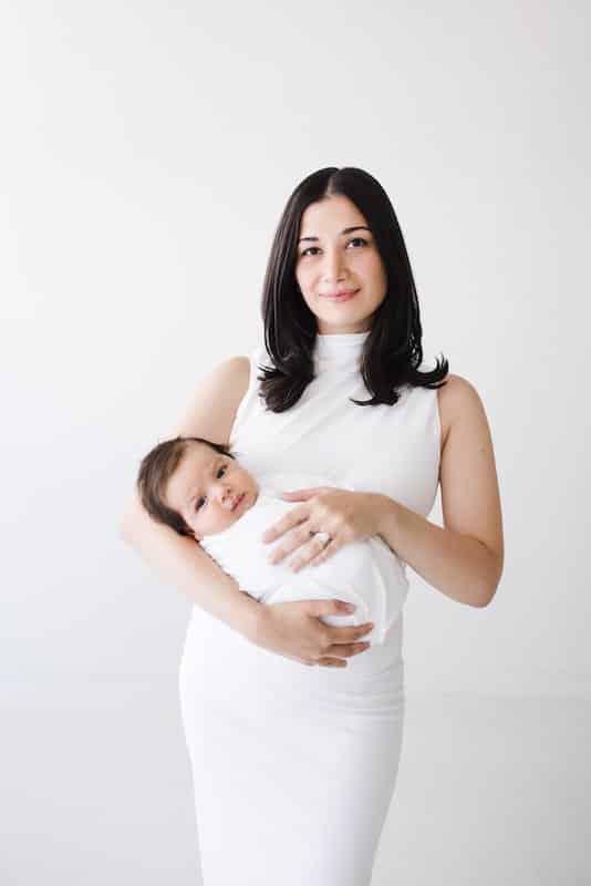 Woman in a white dress holds a swaddled baby against a plain white background.