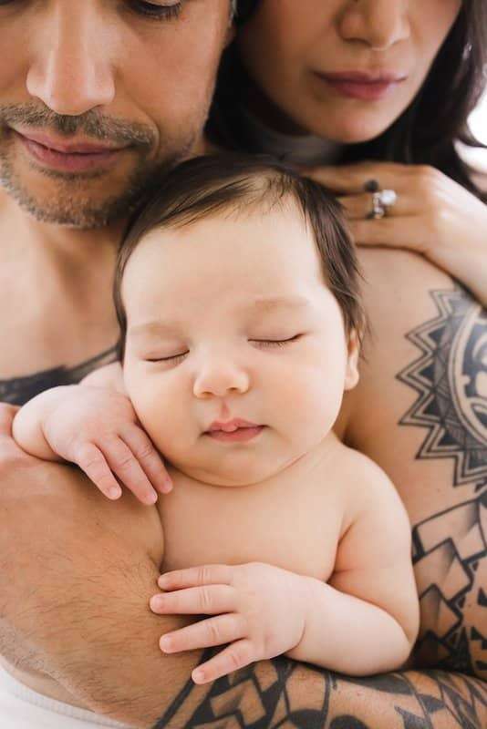 A baby sleeps peacefully in the tattooed arms of two adults, who are close together and partially visible in the background.