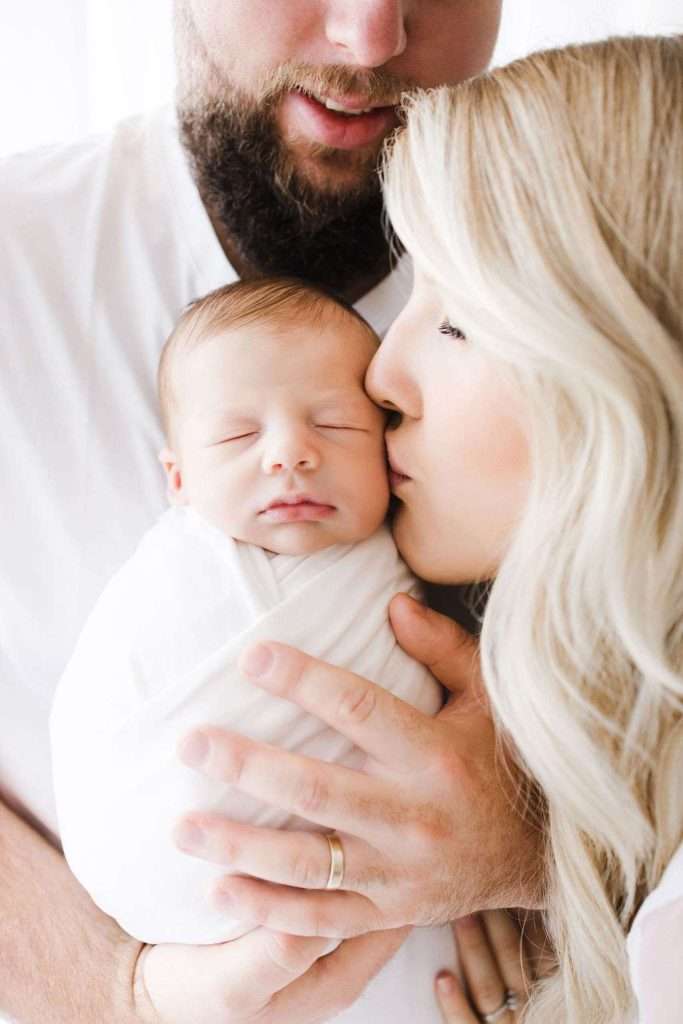 Woman kisses a swaddled sleeping baby held by a man with a beard. All wear white and are closely positioned together, suggesting a family portrait.