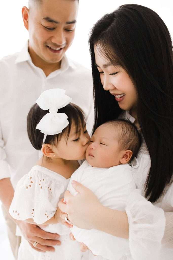A woman holds a swaddled newborn while a young girl with a white bow kisses the baby, and a man stands beside them, all dressed in white.