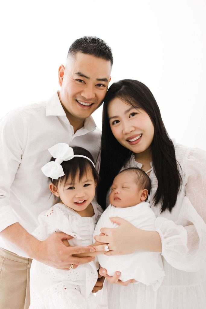 A family of four poses together, with the father and mother standing behind their young daughter and a newborn baby wrapped in a white blanket. All are dressed in light-colored clothing.