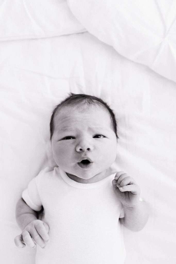 Newborn baby lying on a white blanket, wearing a white onesie, looking up with a slightly open mouth and raised eyebrows.