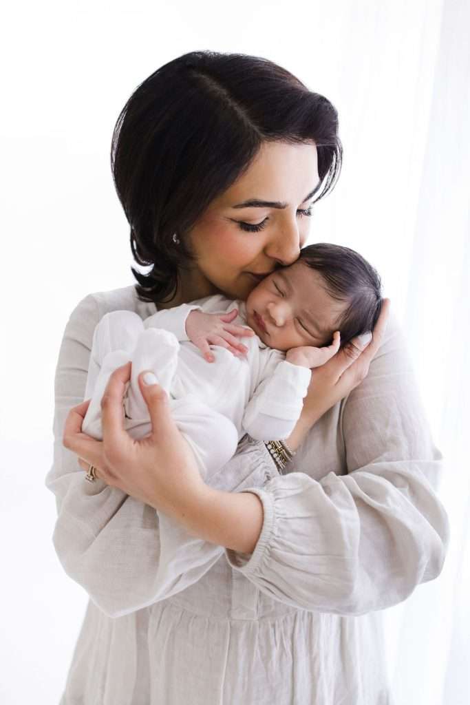 Woman in a light blouse holds and kisses a sleeping newborn dressed in white, both standing against a bright, white background.