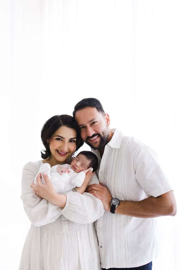 A woman and a man stand close together, smiling, as the woman holds a sleeping newborn in her arms. All three are dressed in white against a bright, light background.