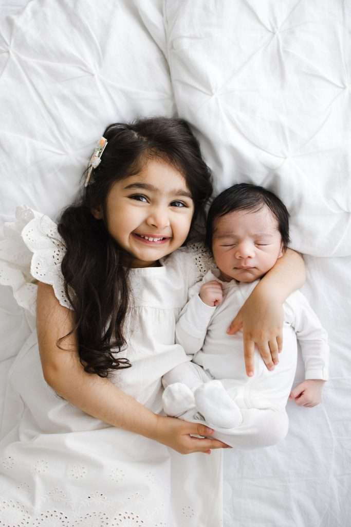 Young girl smiling and hugging a sleeping baby, both dressed in white, lying on a white bed.