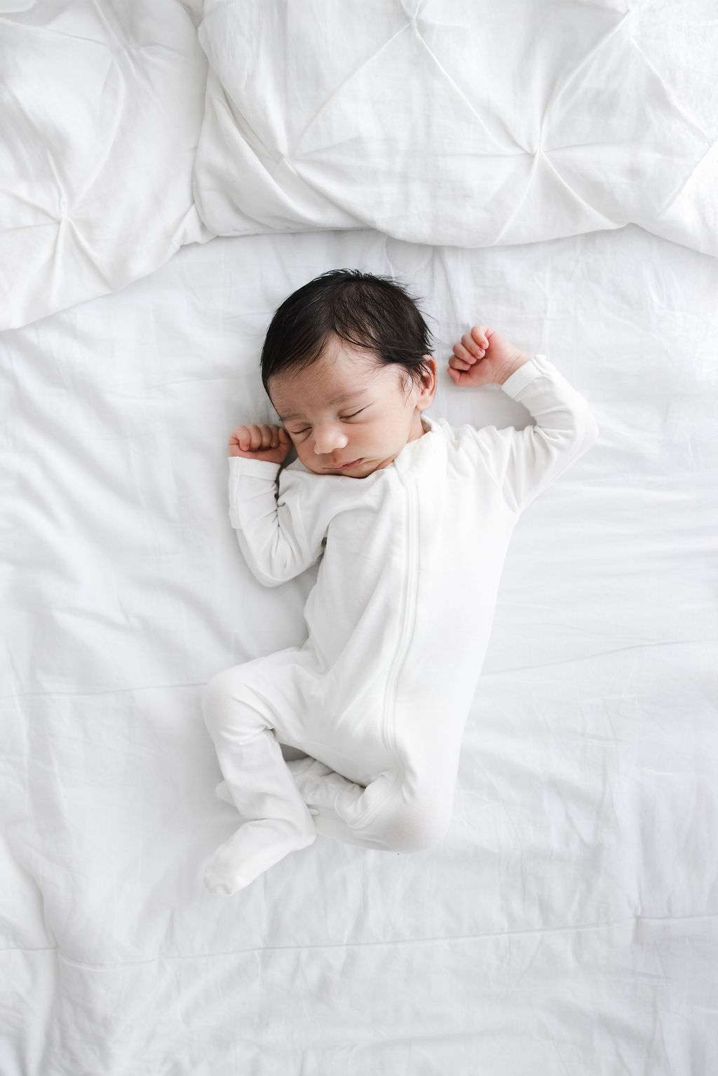 A newborn baby in a white onesie sleeps on a white bed with arms raised and eyes closed.