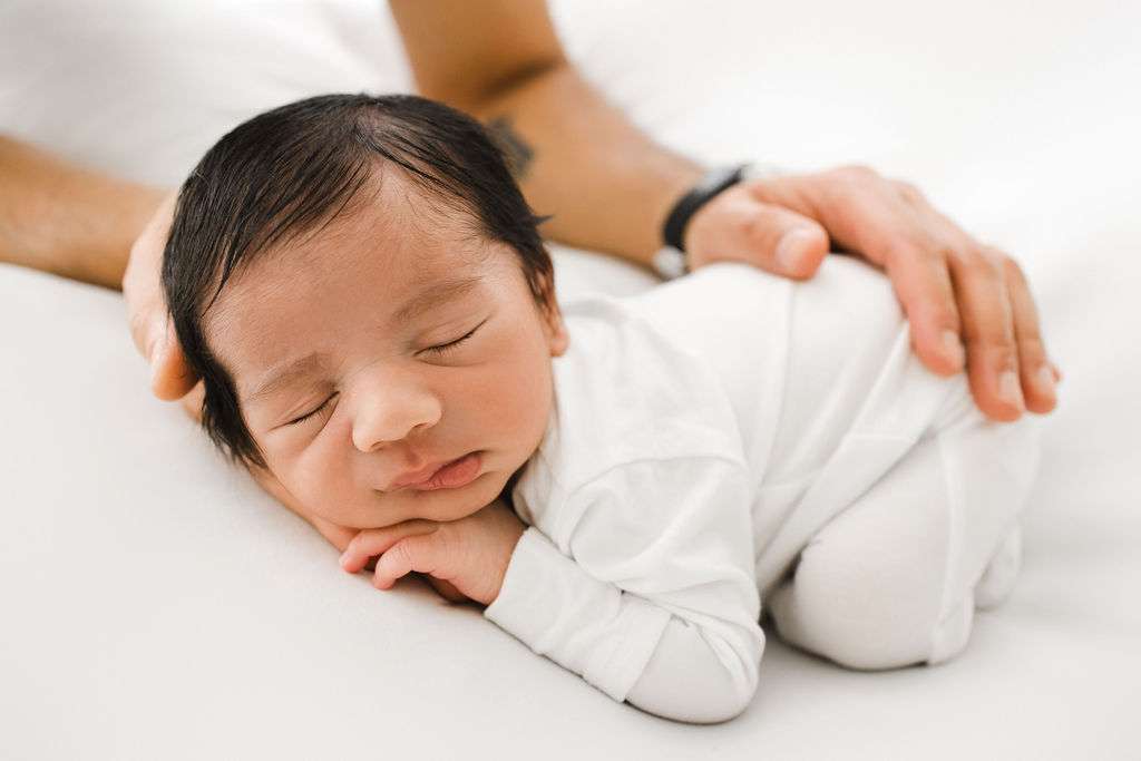 A newborn baby in white clothing sleeps on a white surface, with an adult hand gently resting on the baby's back for support.