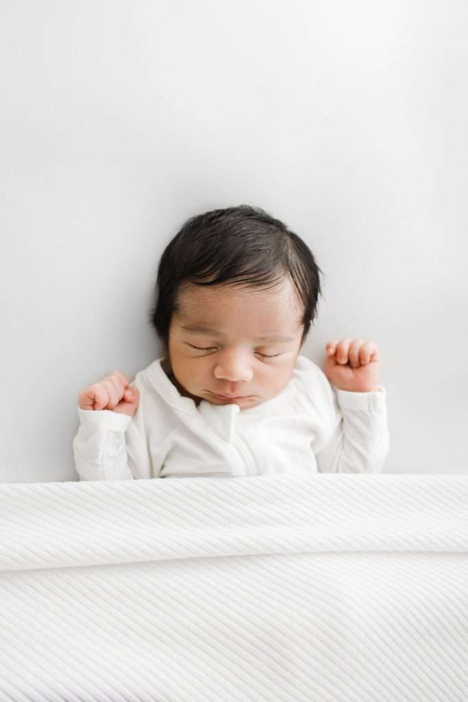 Newborn baby with dark hair sleeping on a white surface, dressed in a white outfit and partially covered with a white blanket.