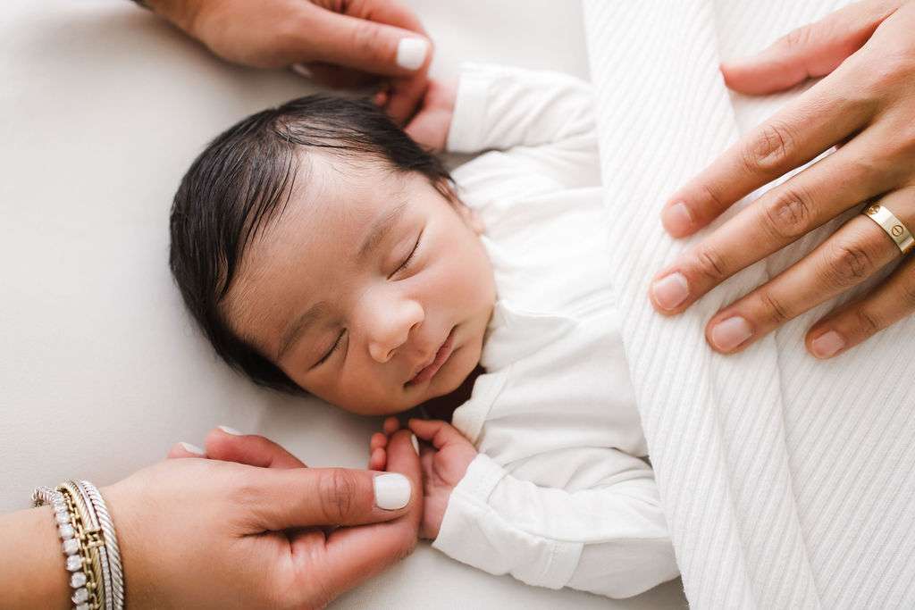 A newborn baby sleeps on a white bed, swaddled in a blanket, with two adult hands gently touching and adjusting the blanket.
