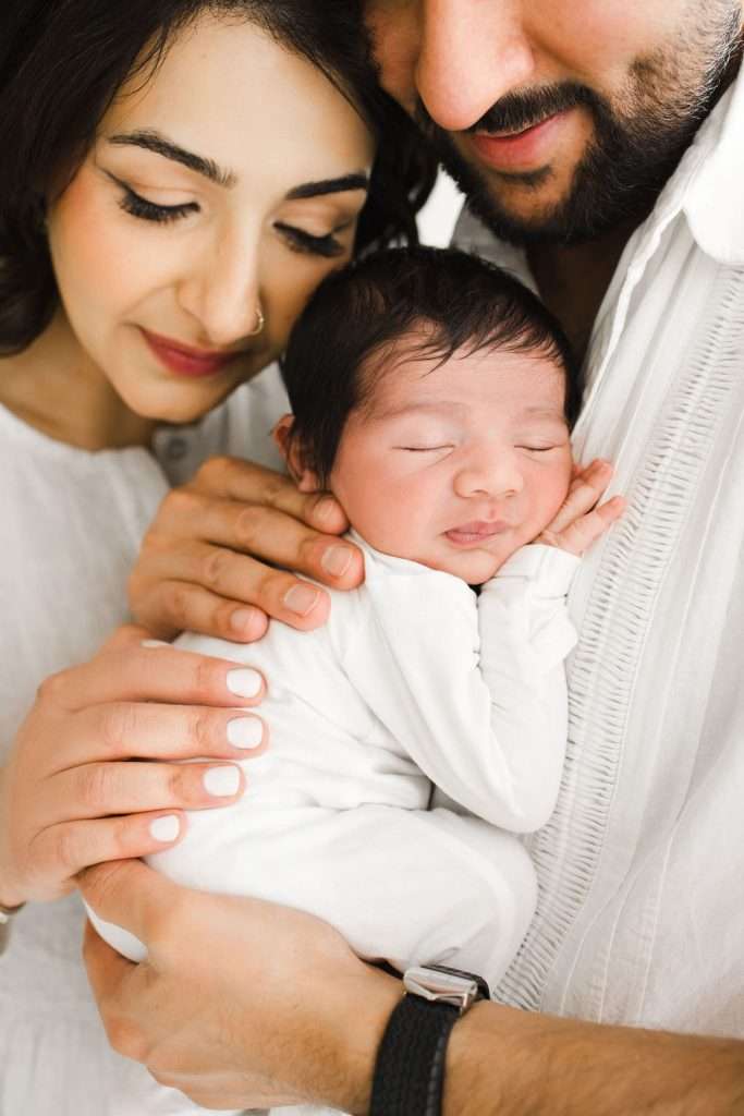 A close-up of two adults holding a sleeping newborn baby, all dressed in white, with gentle expressions and soft lighting.