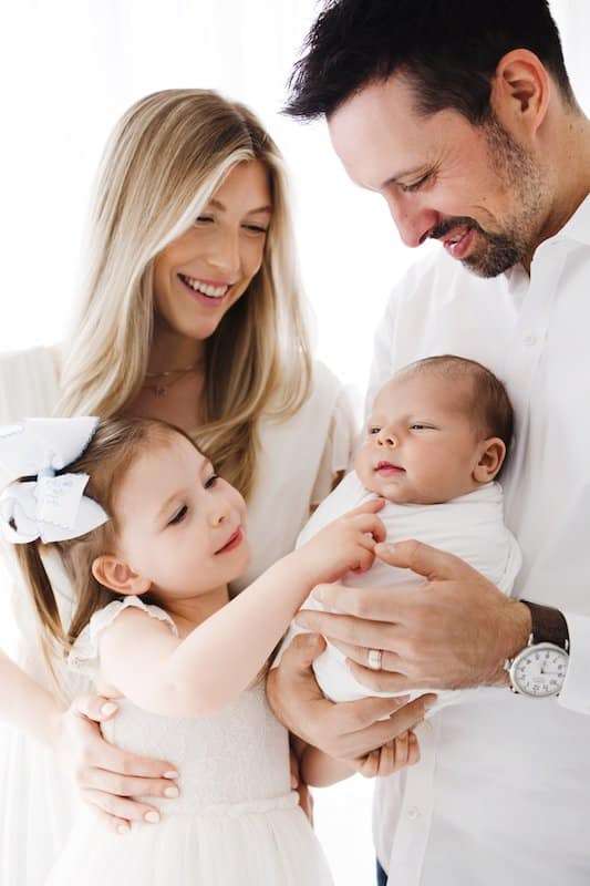 A smiling family of four, with a woman, a man holding a baby, and a young girl gently touching the baby, all dressed in white.