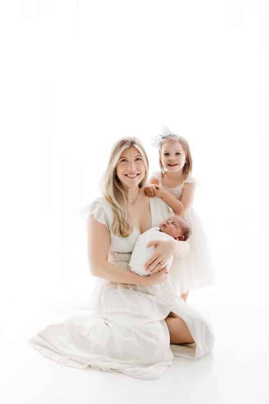 Woman sitting on the floor in a white dress holds a sleeping baby; a young girl in a white dress stands beside her, smiling.