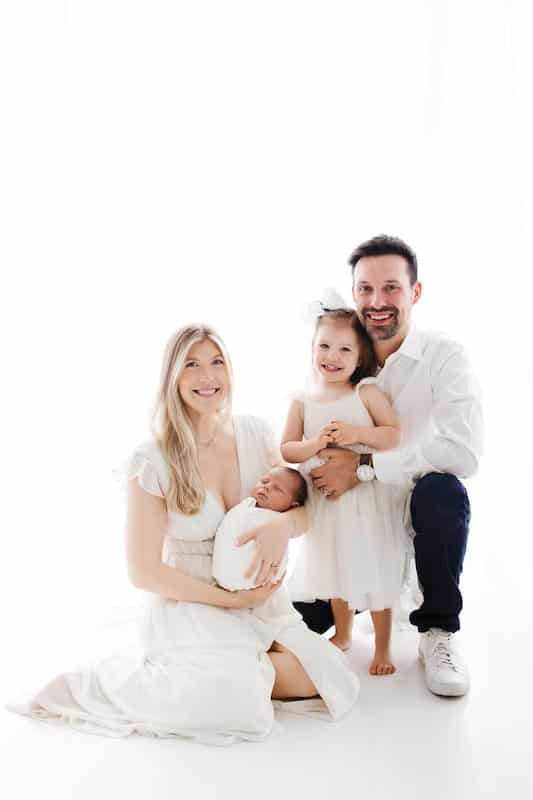 A family of four poses in white clothing against a white background; a woman holds a swaddled baby, a man kneels beside a young girl standing and smiling.