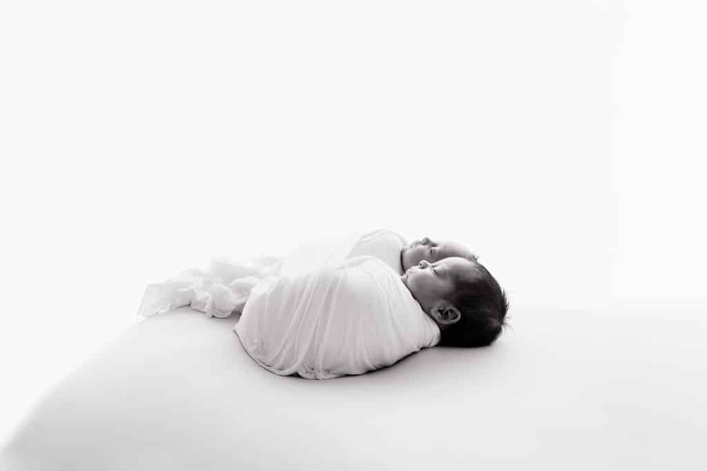 Two newborn babies are wrapped in white blankets and lying side by side on a plain, bright background.