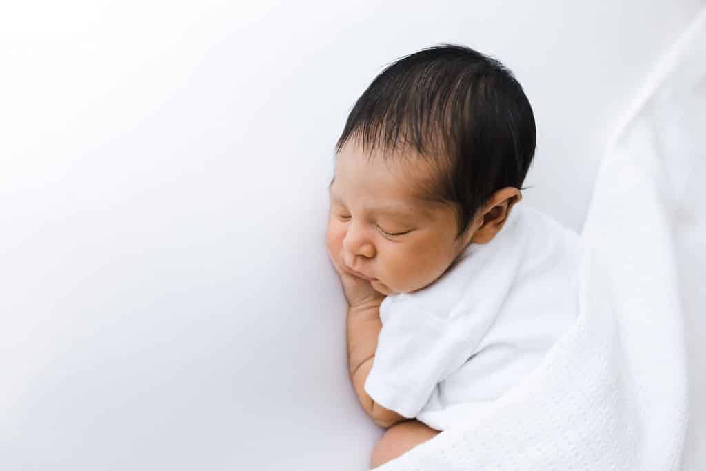 Newborn baby sleeping on its side, wrapped in a white blanket, against a plain white background.