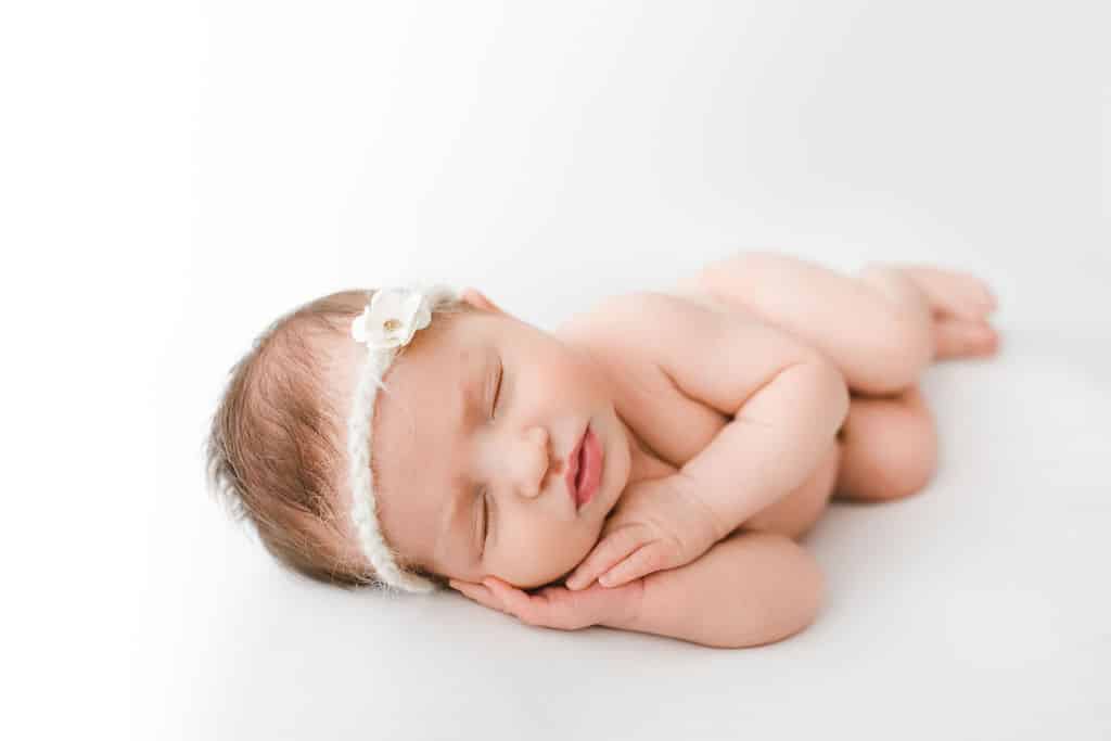Newborn baby sleeping on stomach with hands under cheek, wearing a white headband with a small flower, on a white background.