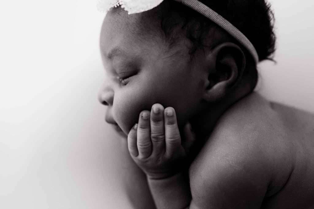 Black and white photo of a baby resting on their side with eyes closed, head propped on one hand, and wearing a headband with a bow.