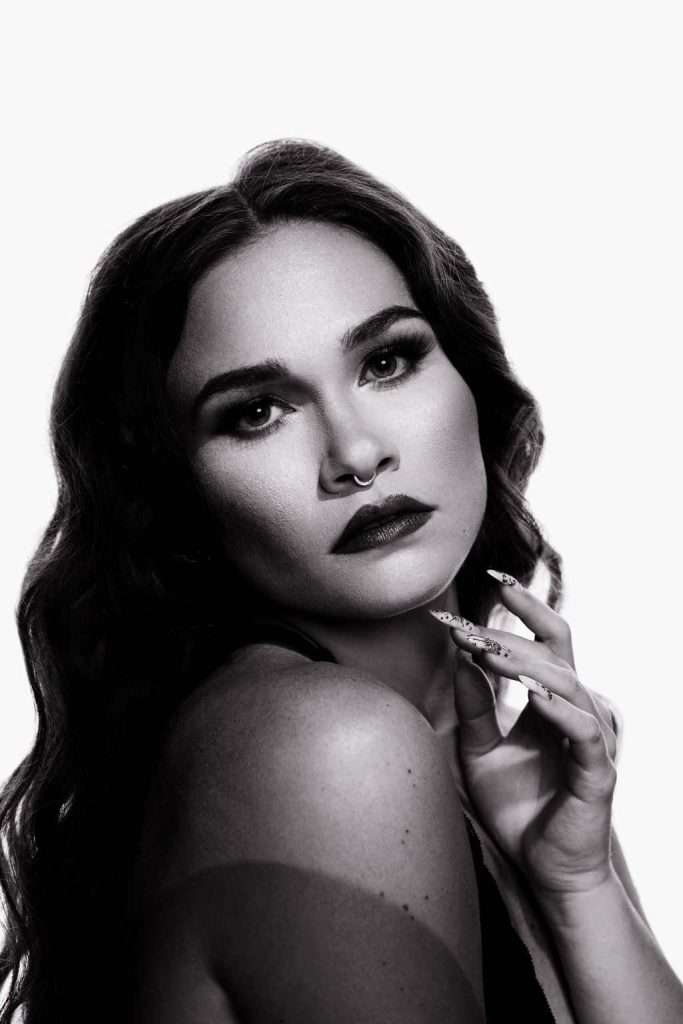 Black and white portrait of a woman with wavy hair, bold makeup, and a septum ring, gazing at the camera while touching her face with manicured nails.