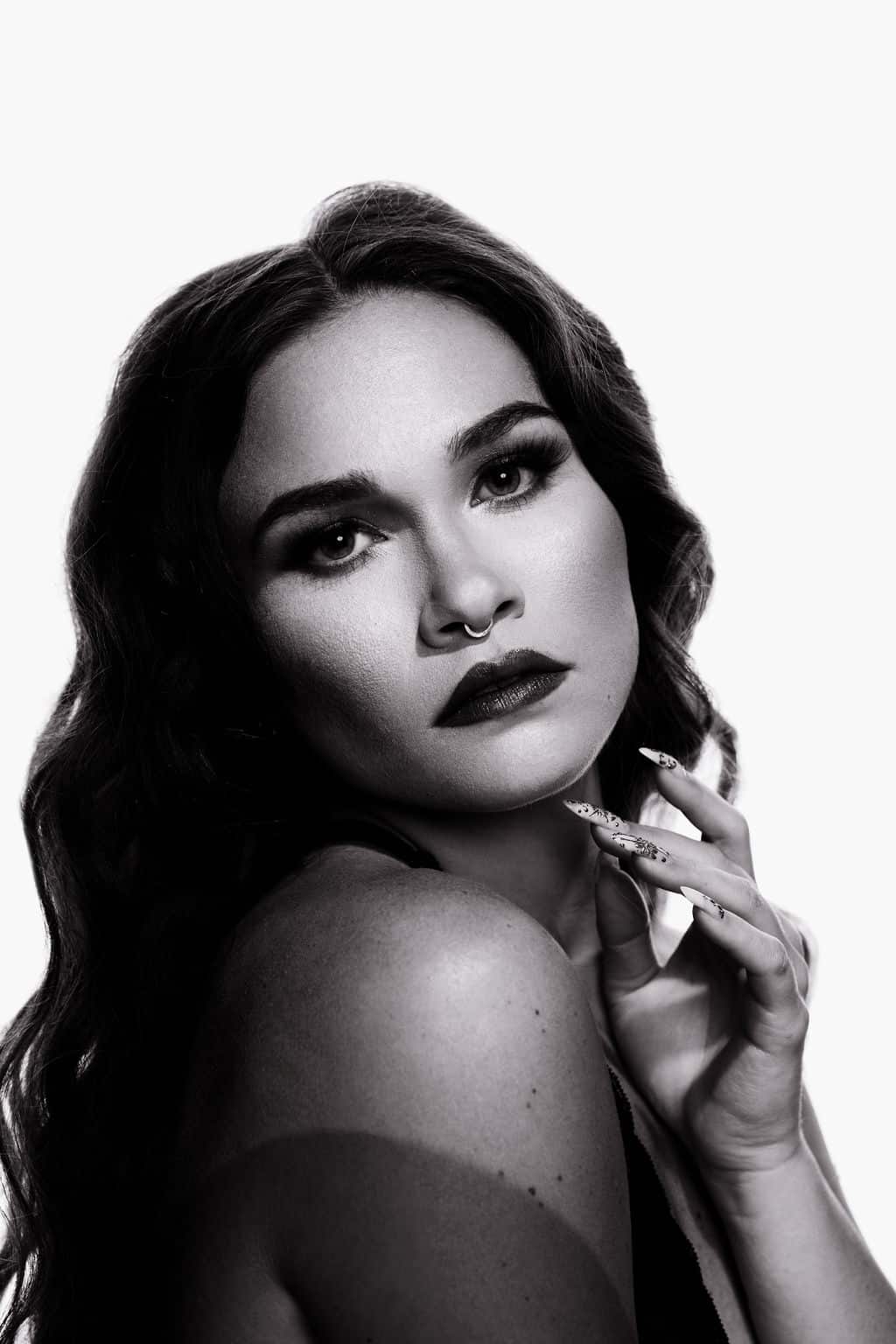 Black and white portrait of a woman with wavy hair, bold makeup, and a septum ring, gazing at the camera while touching her face with manicured nails.