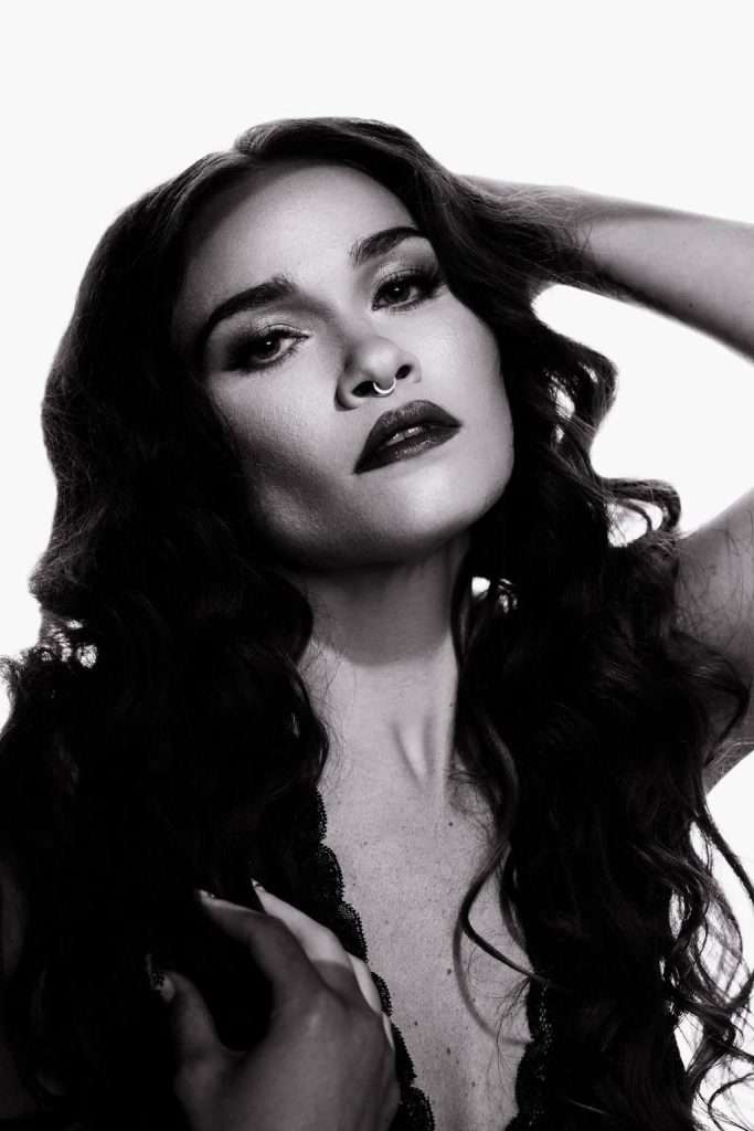 Black and white portrait of a woman with long wavy hair, dark lipstick, and a nose ring, looking at the camera with one hand in her hair.