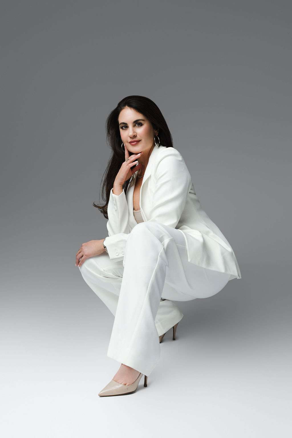 Woman with long dark hair in a white suit squats on the ground, looking at the camera with one hand near her chin. She wears hoop earrings and beige heels against a gray background.