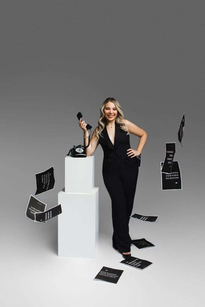 A woman in a black jumpsuit stands by a white pedestal holding a phone, with black cards featuring white text scattered and floating around her, against a plain gray background.