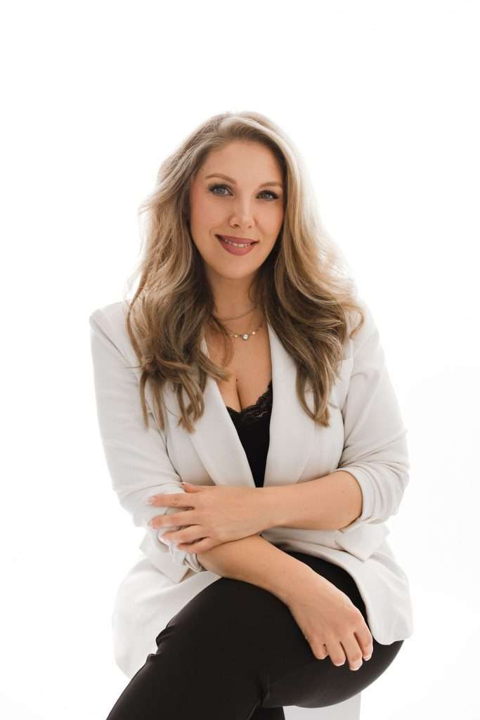 Woman with long wavy hair wearing a white blazer and black pants sits on a stool against a plain white background, smiling at the camera.