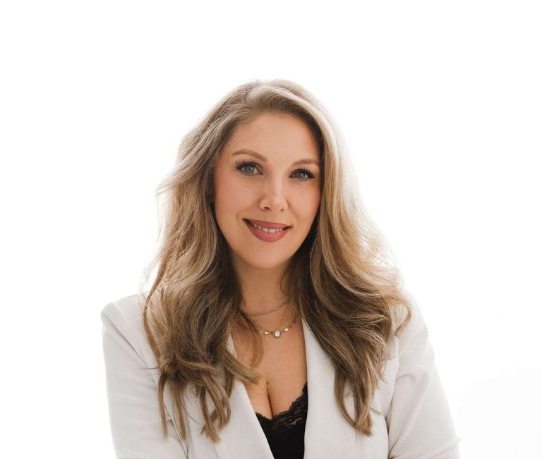 Woman with long wavy hair wearing a white blazer and black pants sits on a stool against a plain white background, smiling at the camera.
