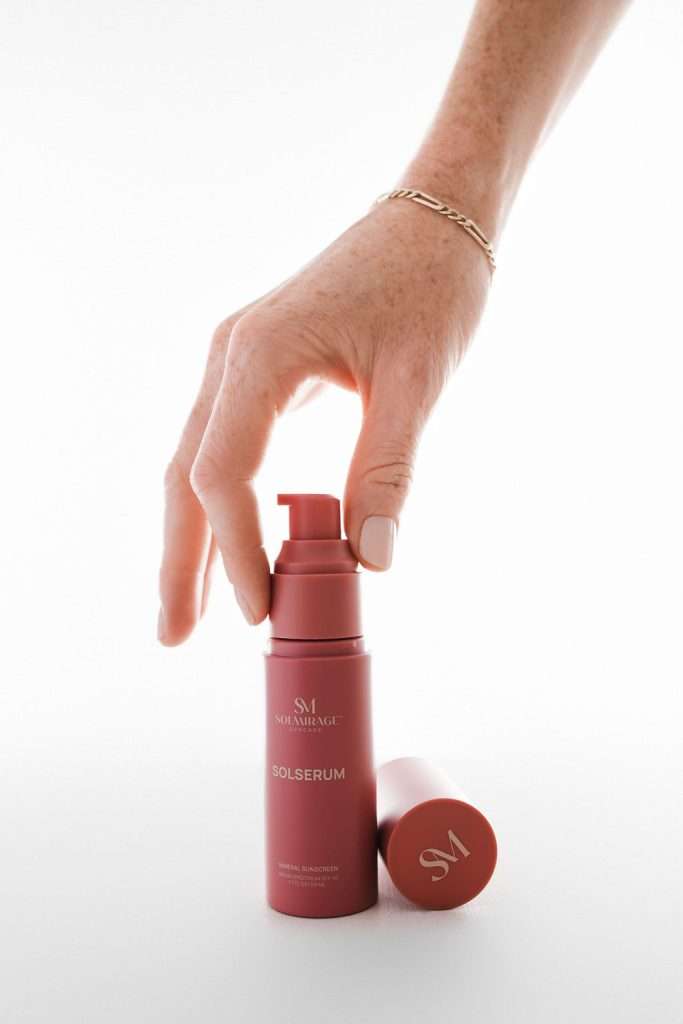 A hand with a gold bracelet holds a pink bottle labeled "SM MARÉAGE SOLSERUM" next to its matching cap, against a white background.