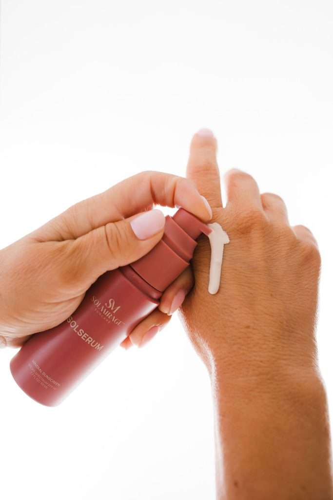A person dispenses white serum from a red bottle onto the back of their hand against a white background.