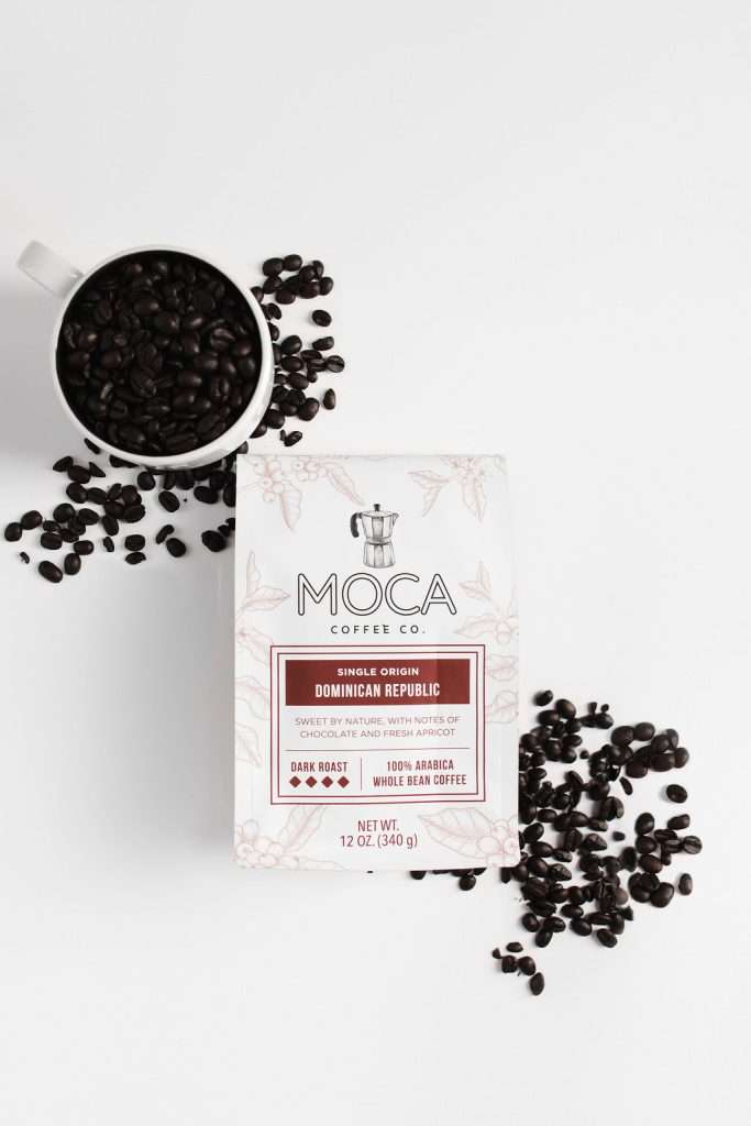 A box of MOCA Dominican Republic single origin coffee sits on a white surface, surrounded by scattered coffee beans and a mug filled with beans.