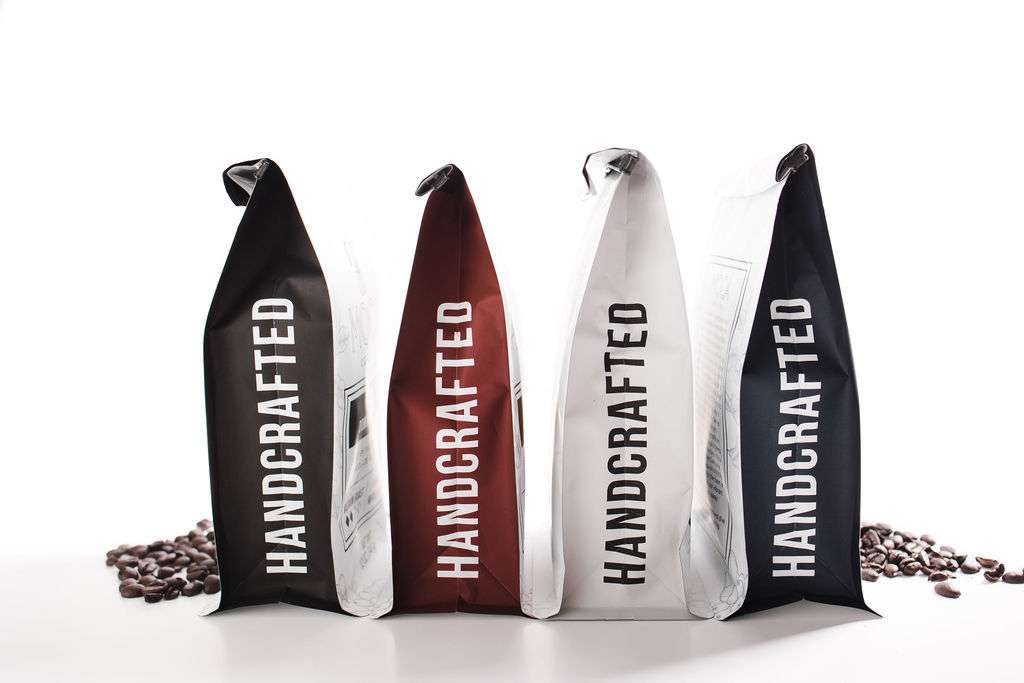 Four upright coffee bags labeled "HANDCRAFTED" in black, brown, and white colors, with coffee beans scattered behind them on a white surface.