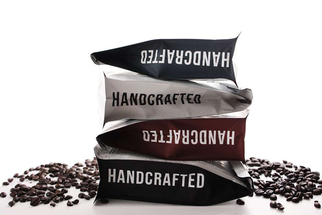 Three bags labeled "HANDCRAFTED" are stacked on each other, surrounded by scattered coffee beans on a white background.
