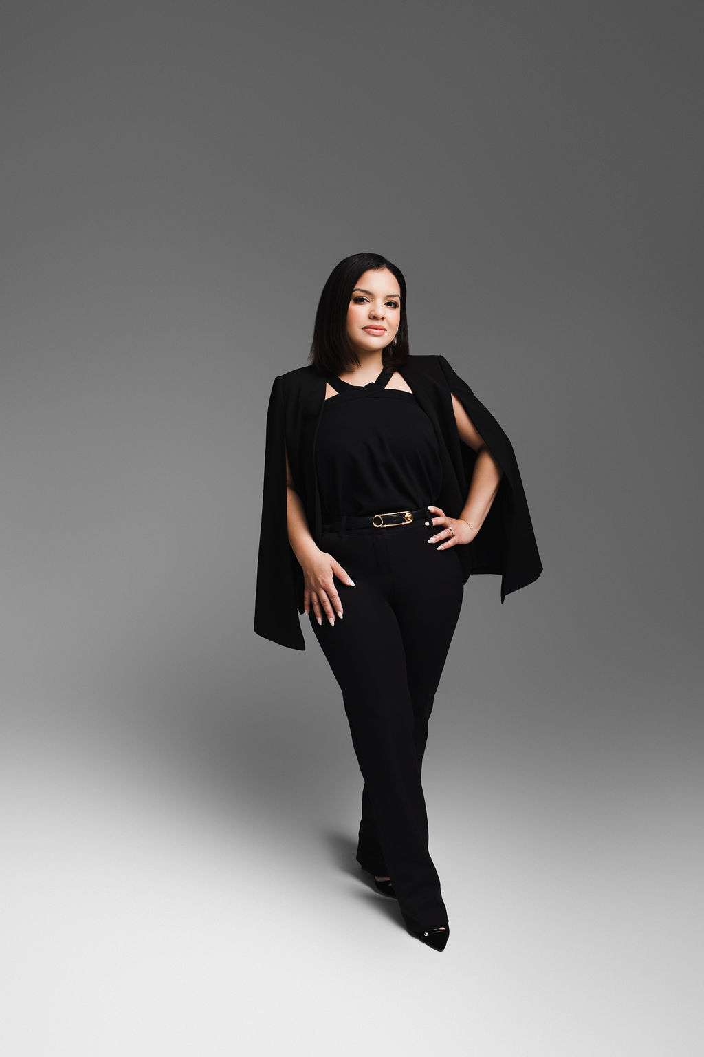 A woman with straight dark hair stands confidently in a black outfit against a gray gradient background, with her hand on her hip and a neutral expression.