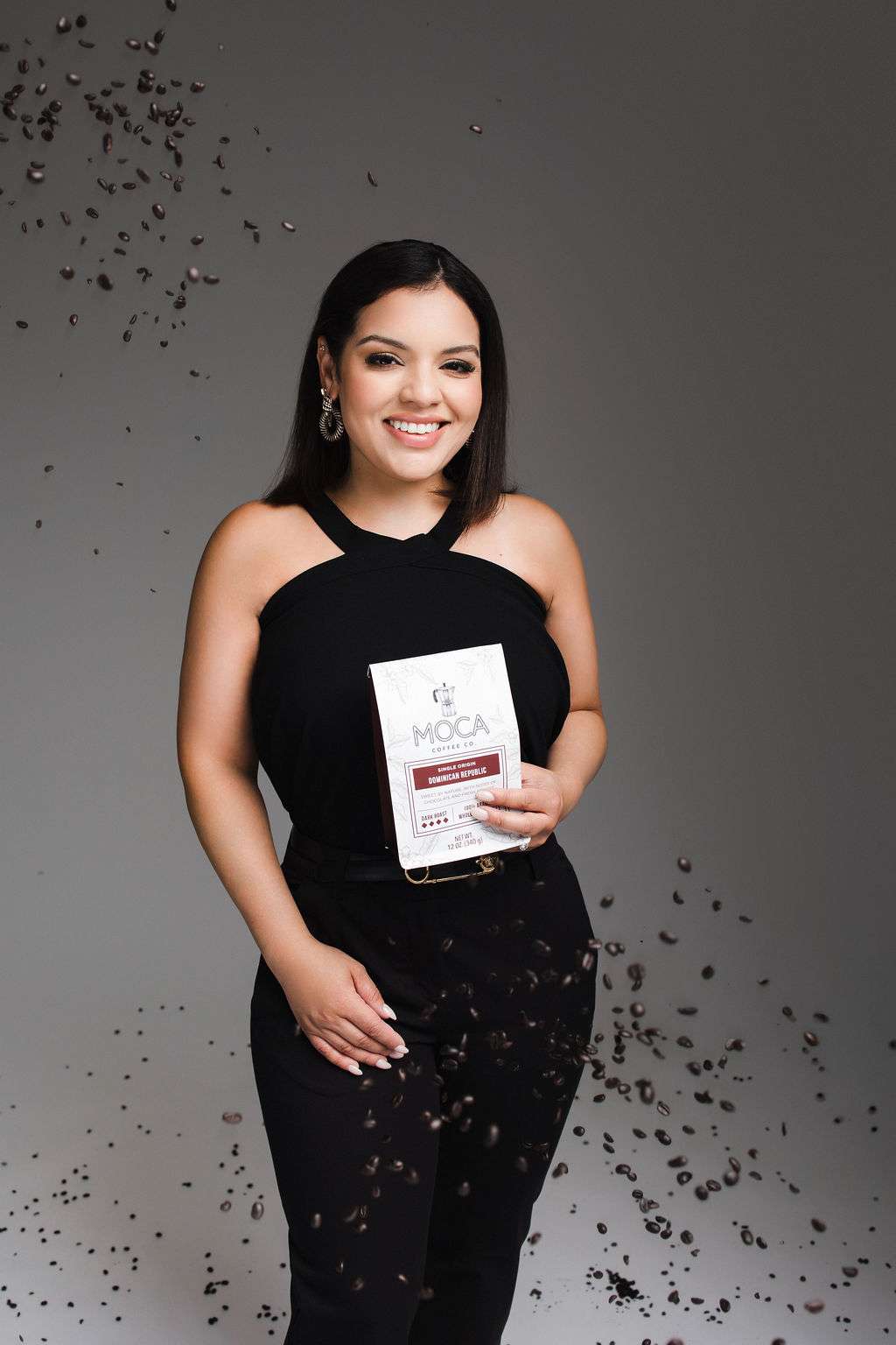 Woman in black outfit holds a bag of coffee labeled "MOCA," surrounded by floating coffee beans against a plain background.