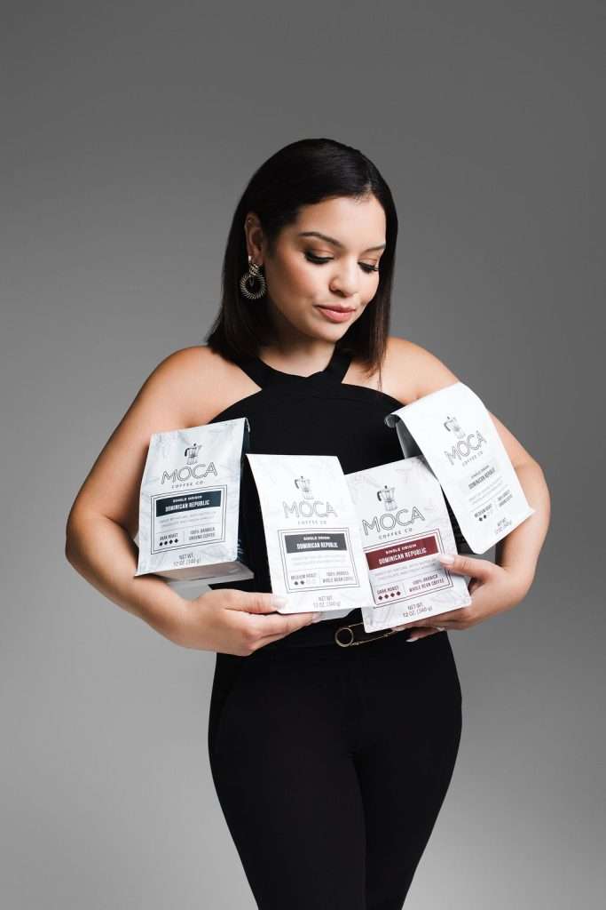 Woman in a black outfit holds four bags of MOCA brand coffee against a plain gray background.