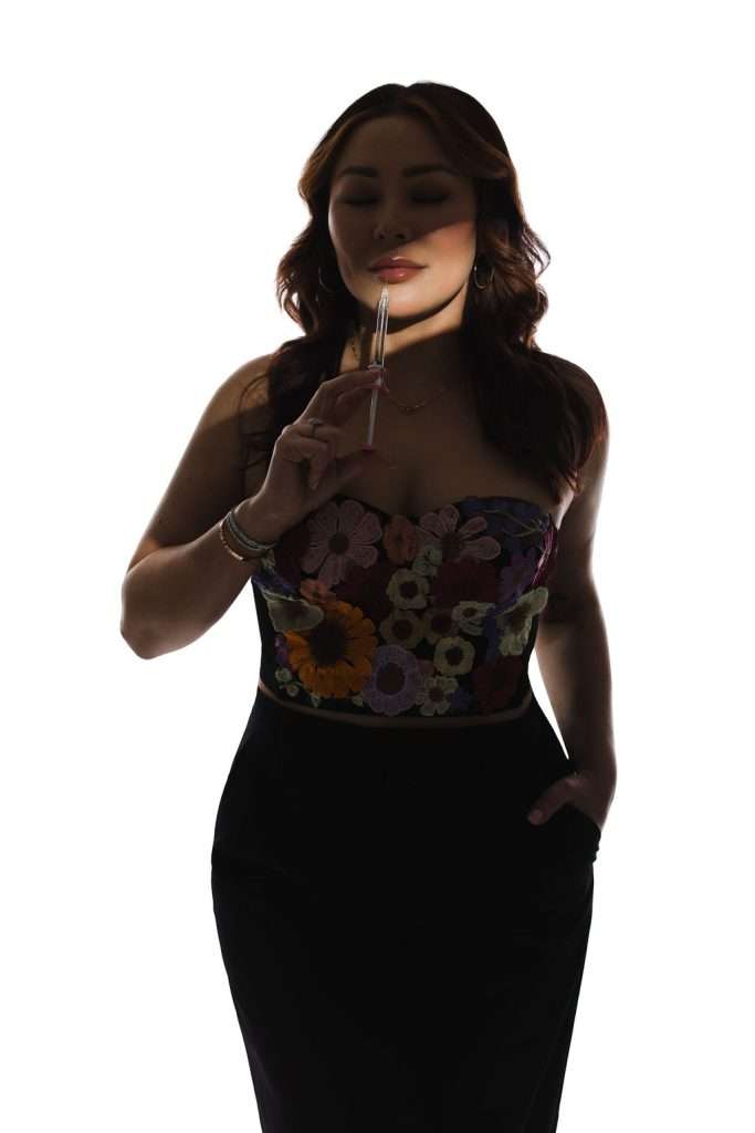 A woman in a floral top and black pants holds a syringe near her lips, standing against a white background with one hand in her pocket.