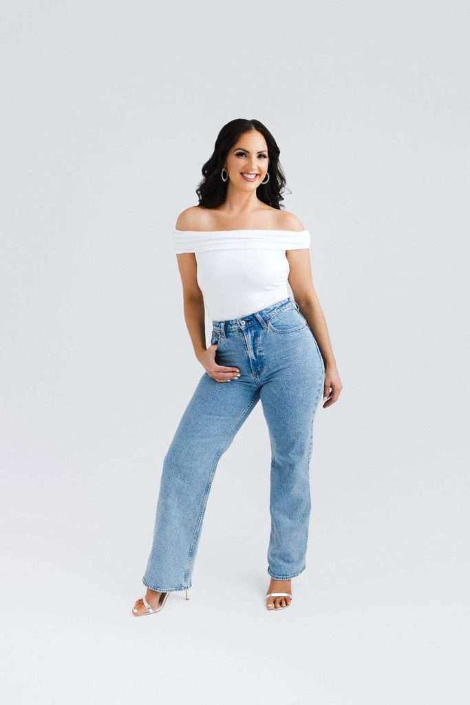 Woman stands smiling, wearing an off-shoulder white top, high-waisted blue jeans, white heels, and hoop earrings against a plain light background.