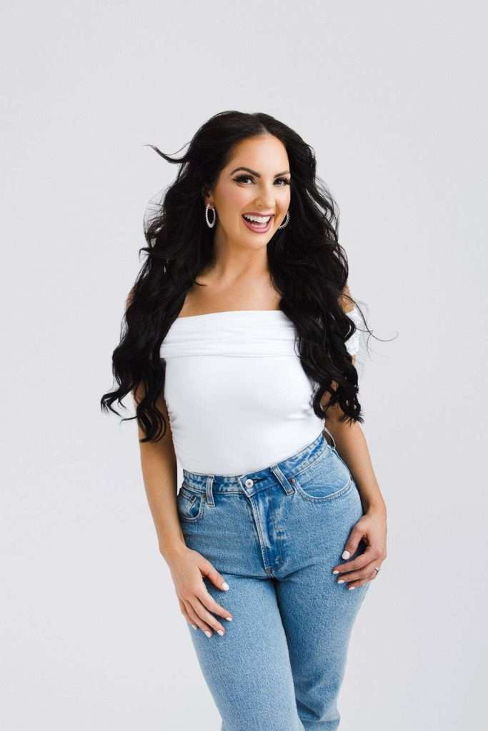 Woman with long dark hair wearing hoop earrings, a white off-shoulder top, and blue jeans stands smiling against a plain light background.