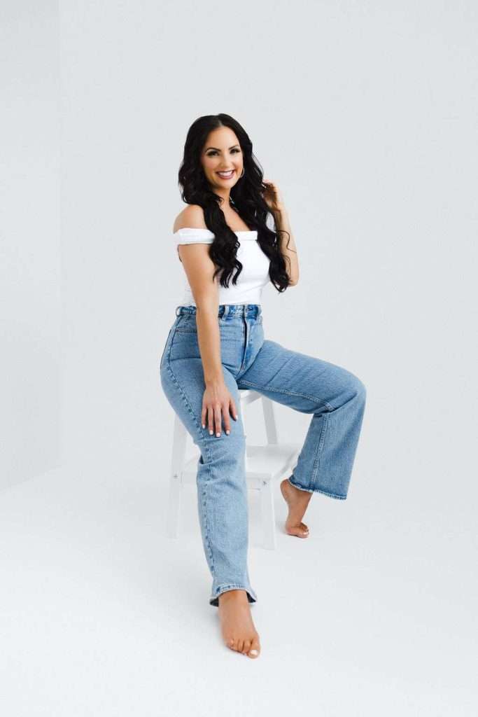 Woman with long dark hair, wearing an off-shoulder white top and blue jeans, sits barefoot on a white stool against a plain light background, smiling at the camera.