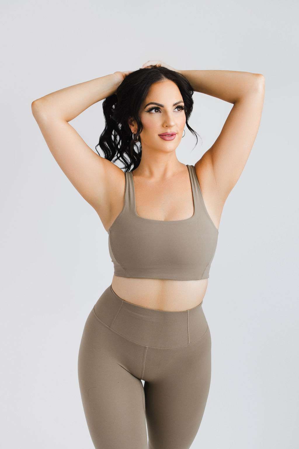 Woman in taupe sports bra and leggings stands with hands on head, looking to the side, against a plain light background.