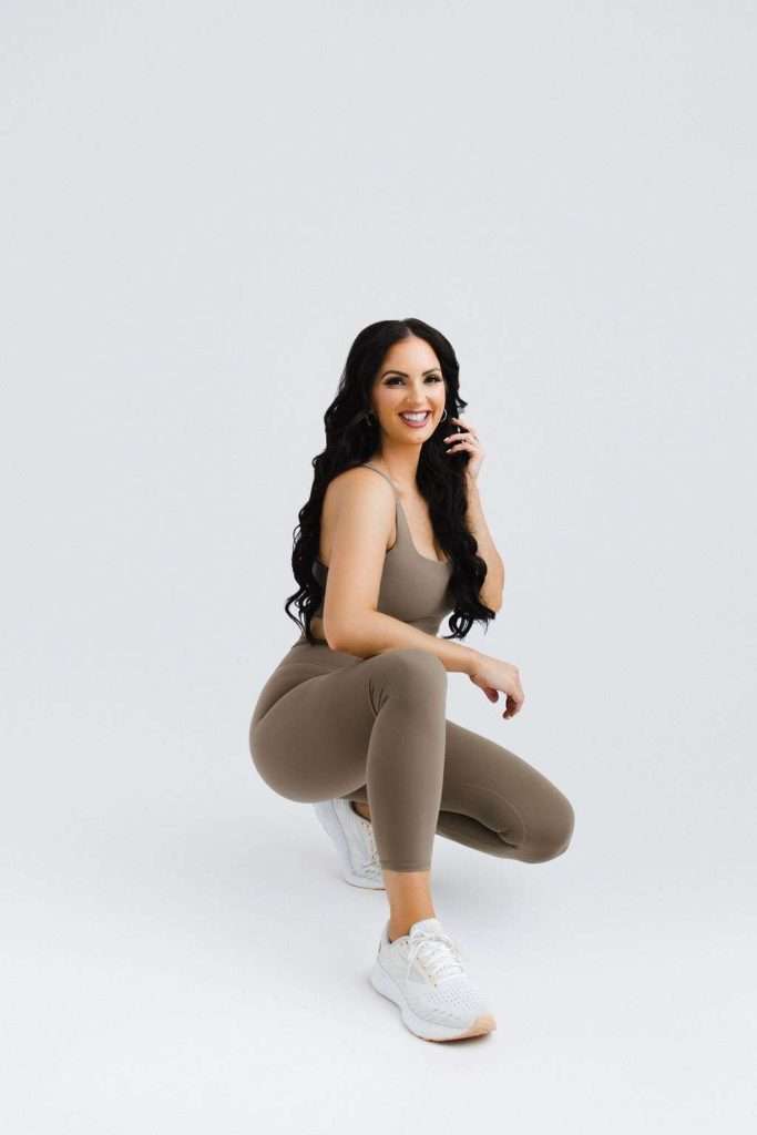 Woman with long dark hair squats on one knee, smiling at the camera, wearing taupe activewear and white sneakers against a plain light background.