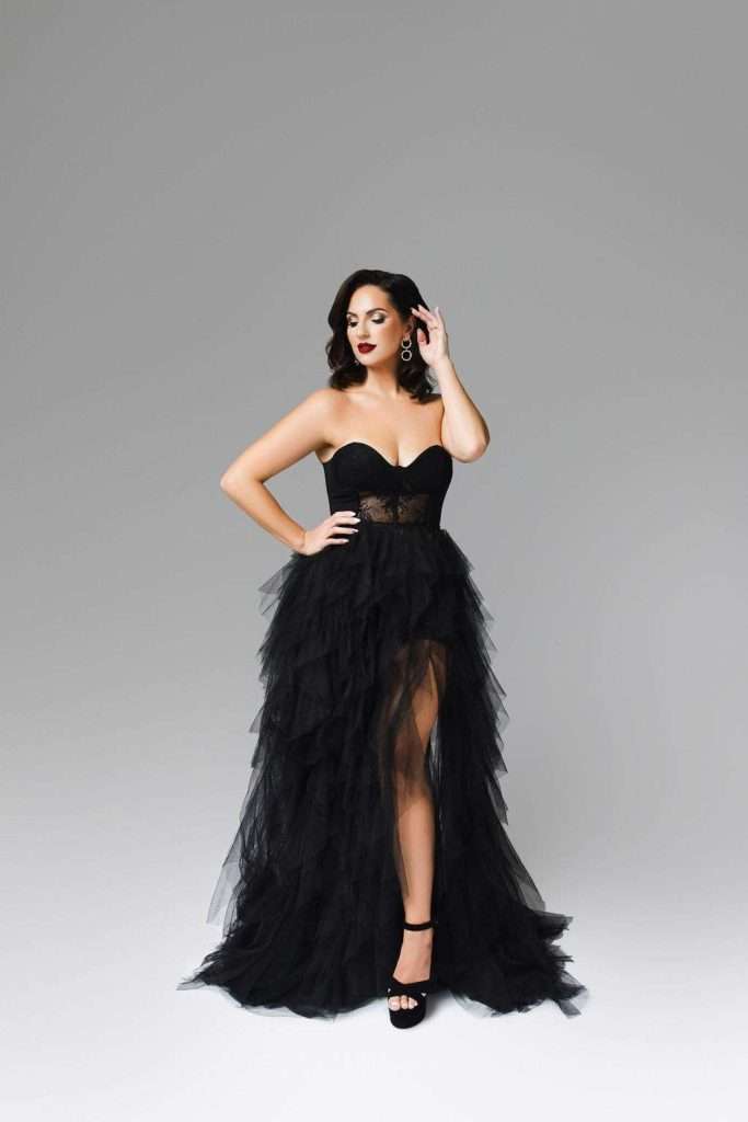 Woman in a strapless black tulle evening gown with a high slit poses against a plain gray background, wearing black heels and statement earrings.