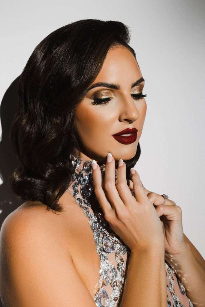 Woman with dark wavy hair and bold makeup poses in a beaded, sleeveless dress, gently touching her face and neck with closed eyes against a neutral background.