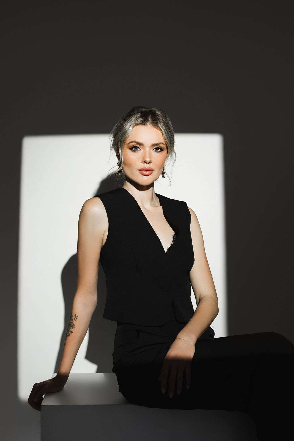 Woman with light hair sits on a white block, wearing a black sleeveless top, with a square of light illuminating her against a dark background.