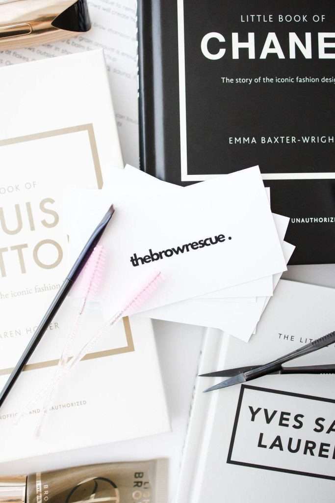 White cards labeled "thebrowrescue" are placed on top of fashion books about Chanel, Louis Vuitton, and Yves Saint Laurent, with two pink eyebrow brushes nearby.