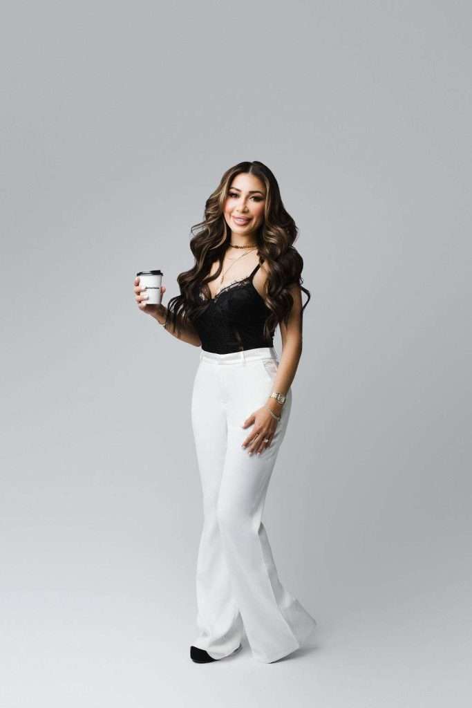 Woman with long wavy hair wearing a black top and white pants stands holding a disposable coffee cup against a plain light gray background.