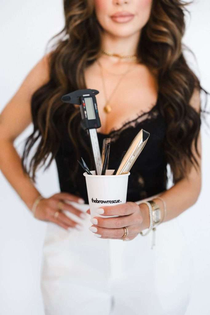 A woman in a black top and white pants holds a cup labeled "thebrowrescue" containing beauty tools and a caliper.