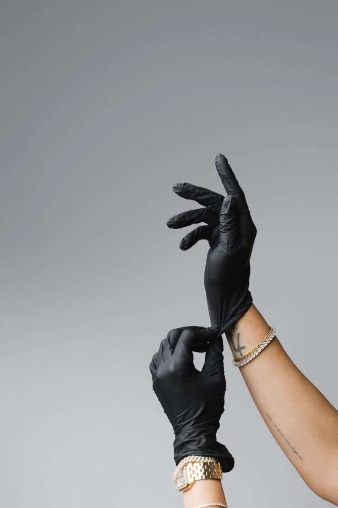 A person wearing a watch and bracelets puts on a pair of black disposable gloves against a plain gray background.