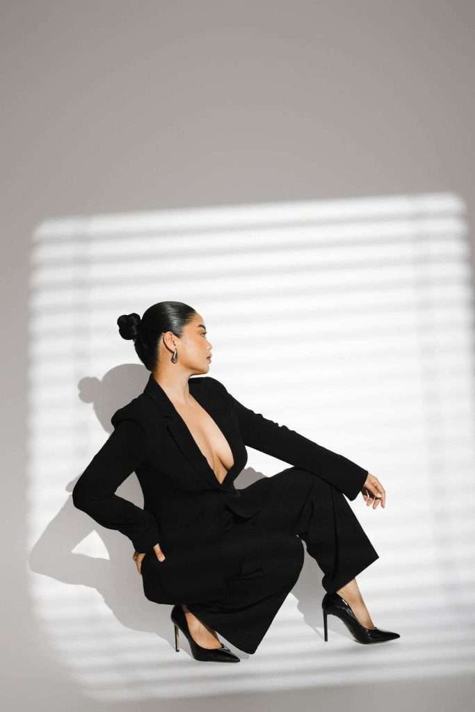 Person in a black suit and heels sits on the floor in profile, with hair in a bun, lit by sunlight streaming through window blinds.